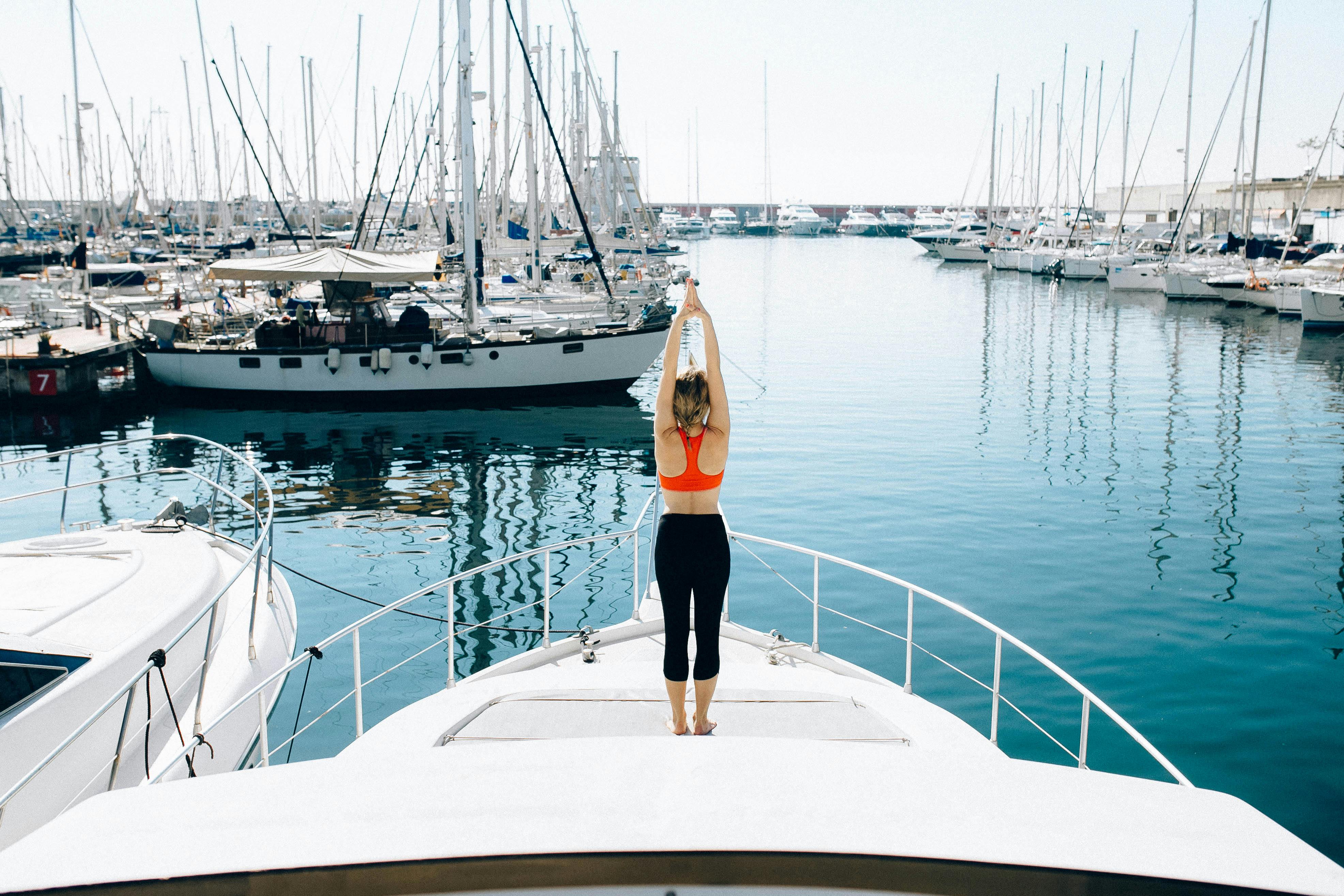 Woman in orange sports bra and black leggings doing yoga stretch on the bow of a white boat in a marina with many sailboats.