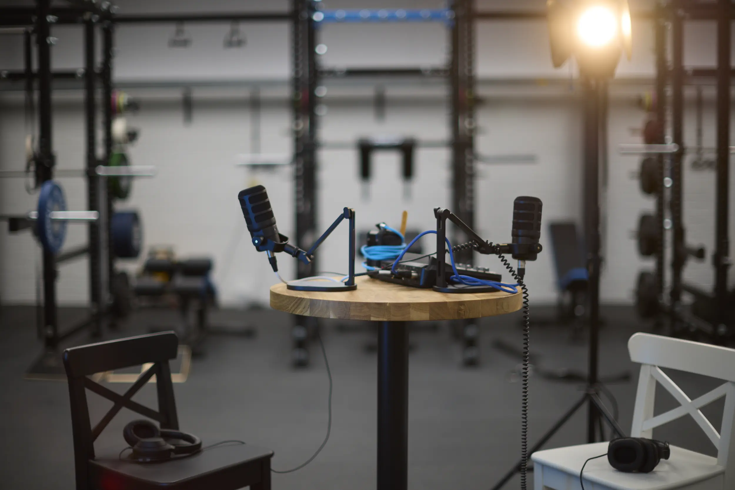 Podcast setup with two microphones on a round wooden table and headphones on two chairs in a gym with weightlifting equipment in the background.