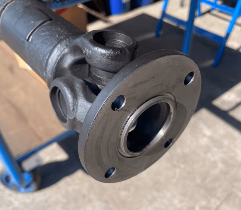 Nahaufnahme einer schwarzen Metallwelle mit Flansch und Schraublöchern. Schatten und blauer Metallständer im Hintergrund.