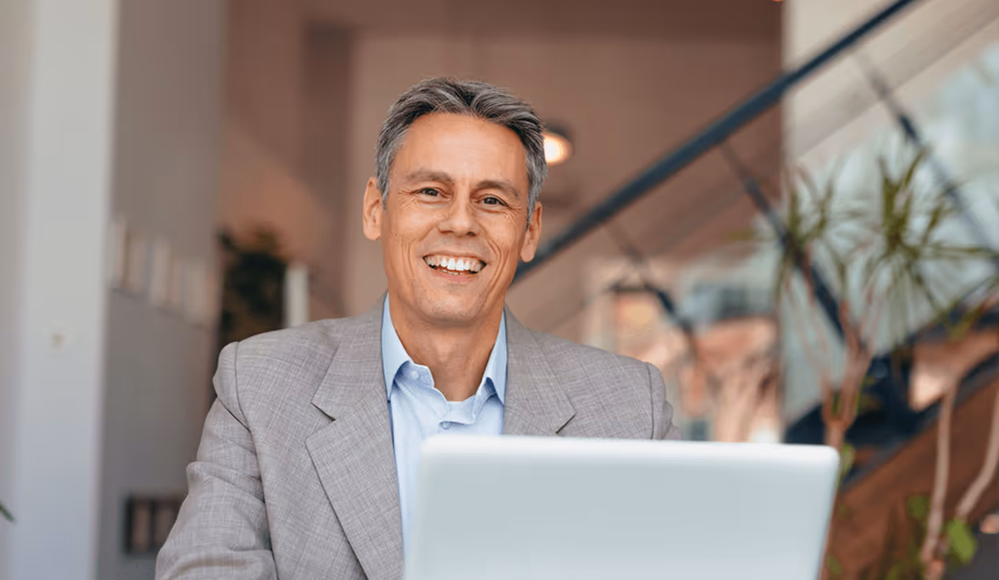 Mann in grauem Anzug lächelt vor Laptop, Treppe im Hintergrund, entspanntes Arbeitsumfeld.
