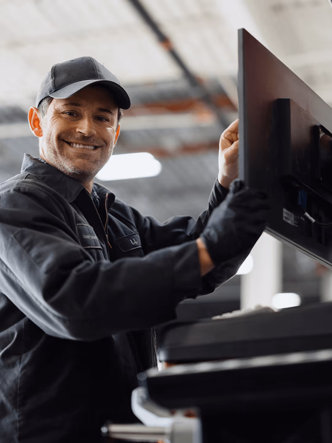 Techniker repariert Monitor in Werkstatt, trägt schwarze Kleidung und Handschuhe, lächelt.