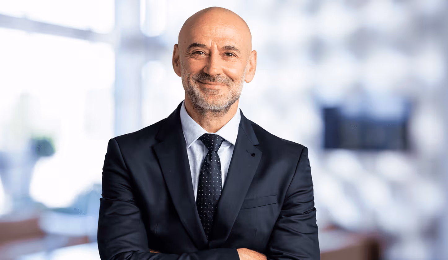 Lächelnder, glatzköpfiger Mann im Anzug mit verschränkten Armen vor verschwommenem Hintergrund im Büro.