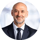 Ein lächelnder glatzköpfiger Mann in Anzug und Krawatte vor verschwommenem Hintergrund.