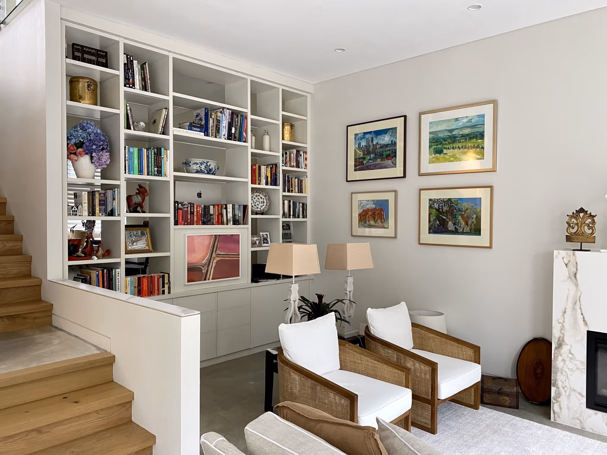 Bright living room with built-in white bookshelves filled with books and decor, wooden stairs on the left, two white cushioned wooden armchairs, framed paintings on the wall, and a marble fireplace on the right.