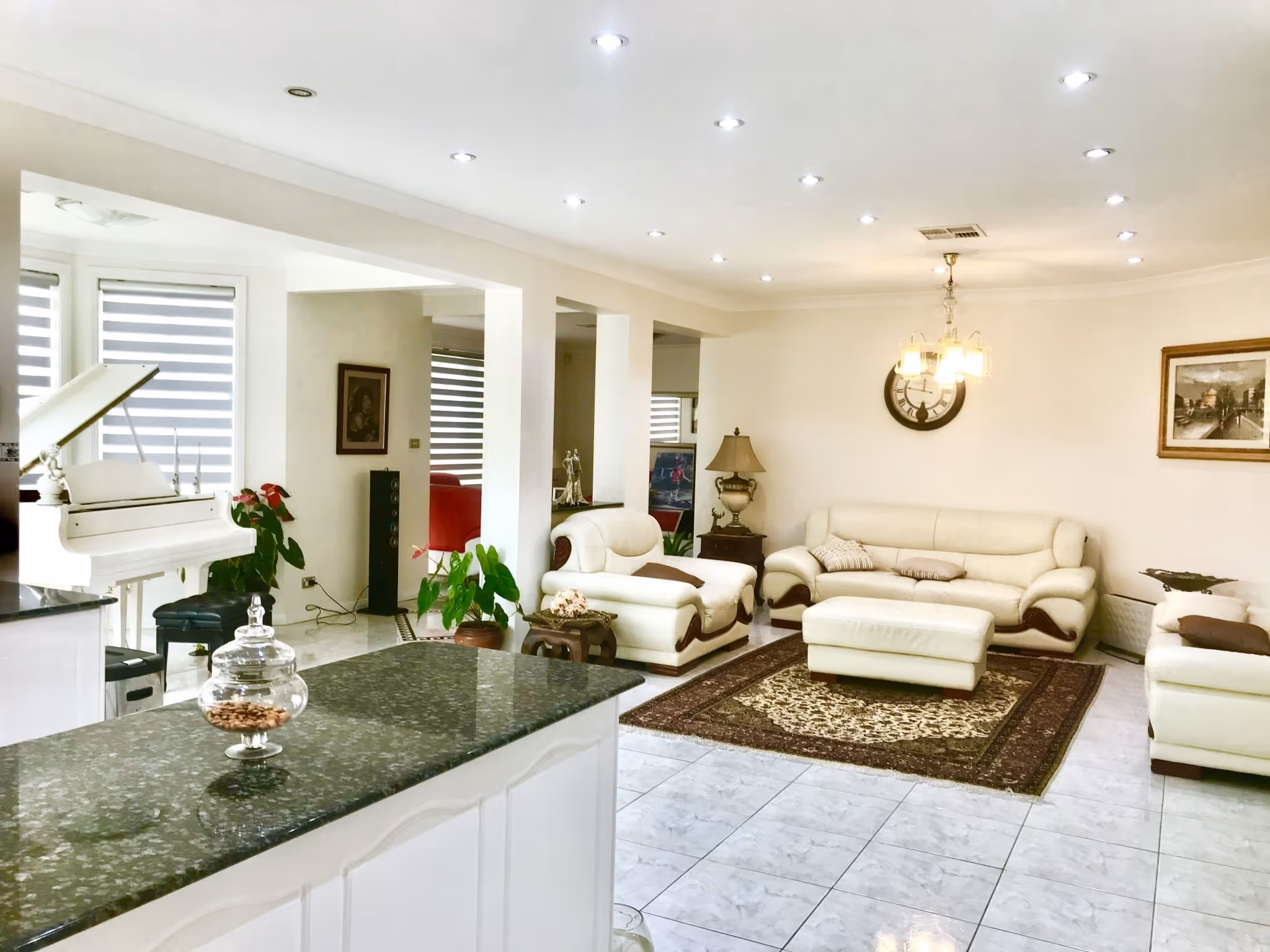 Bright living room with white leather sofas, a patterned rug, wall clock, paintings, and a white grand piano near a kitchen island.