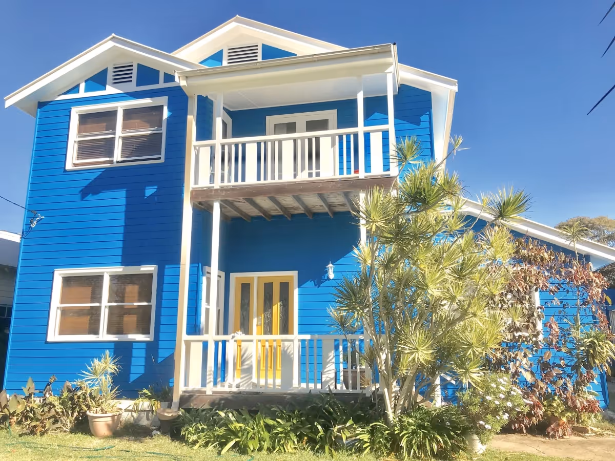 Two-story blue house with white trim, yellow front door, balcony, and surrounding plants under clear blue sky.
