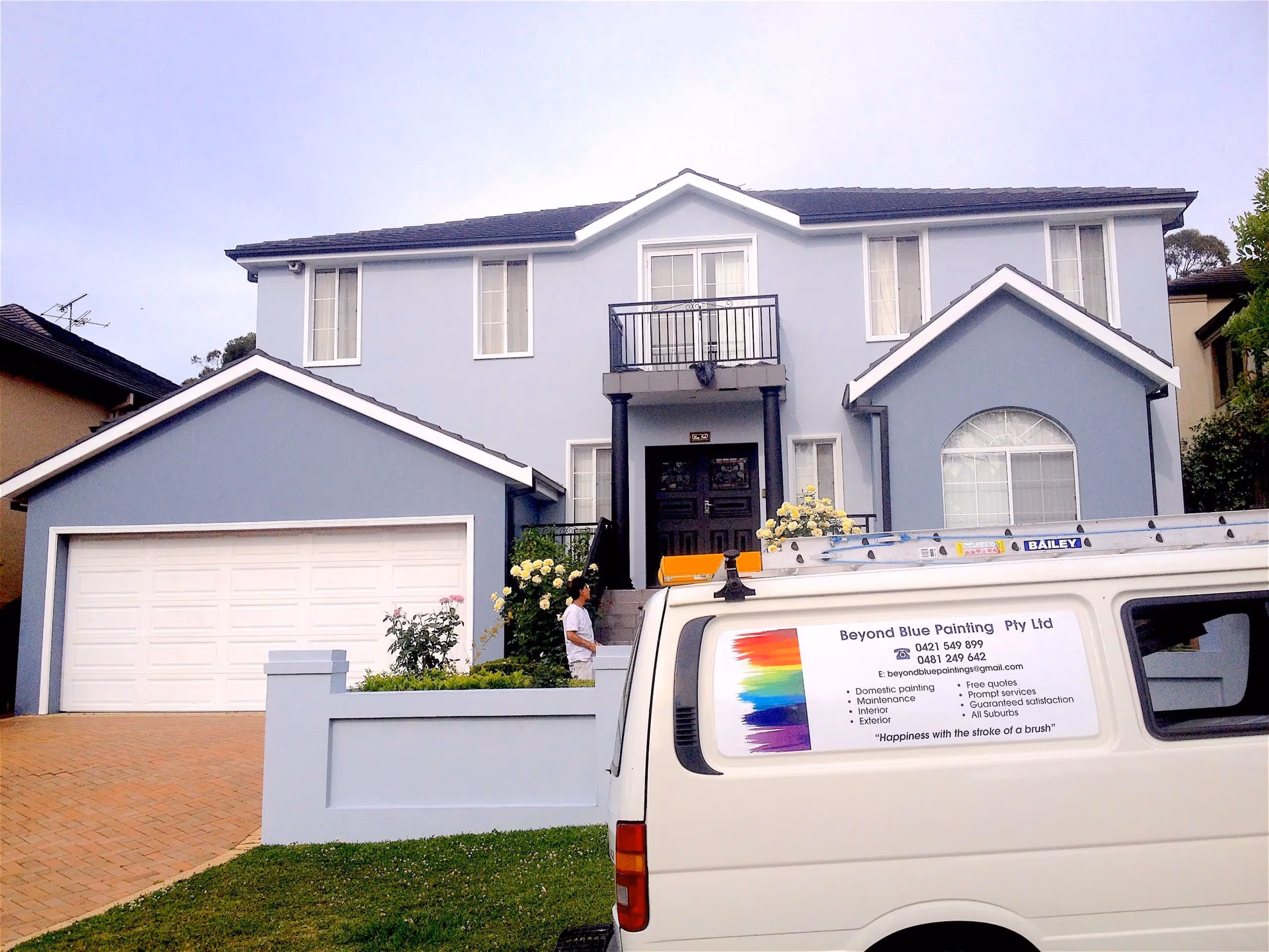 Two-story blue house with white trim and a white garage door, a person standing by the front steps, and a white van with painting company signage parked in the driveway.