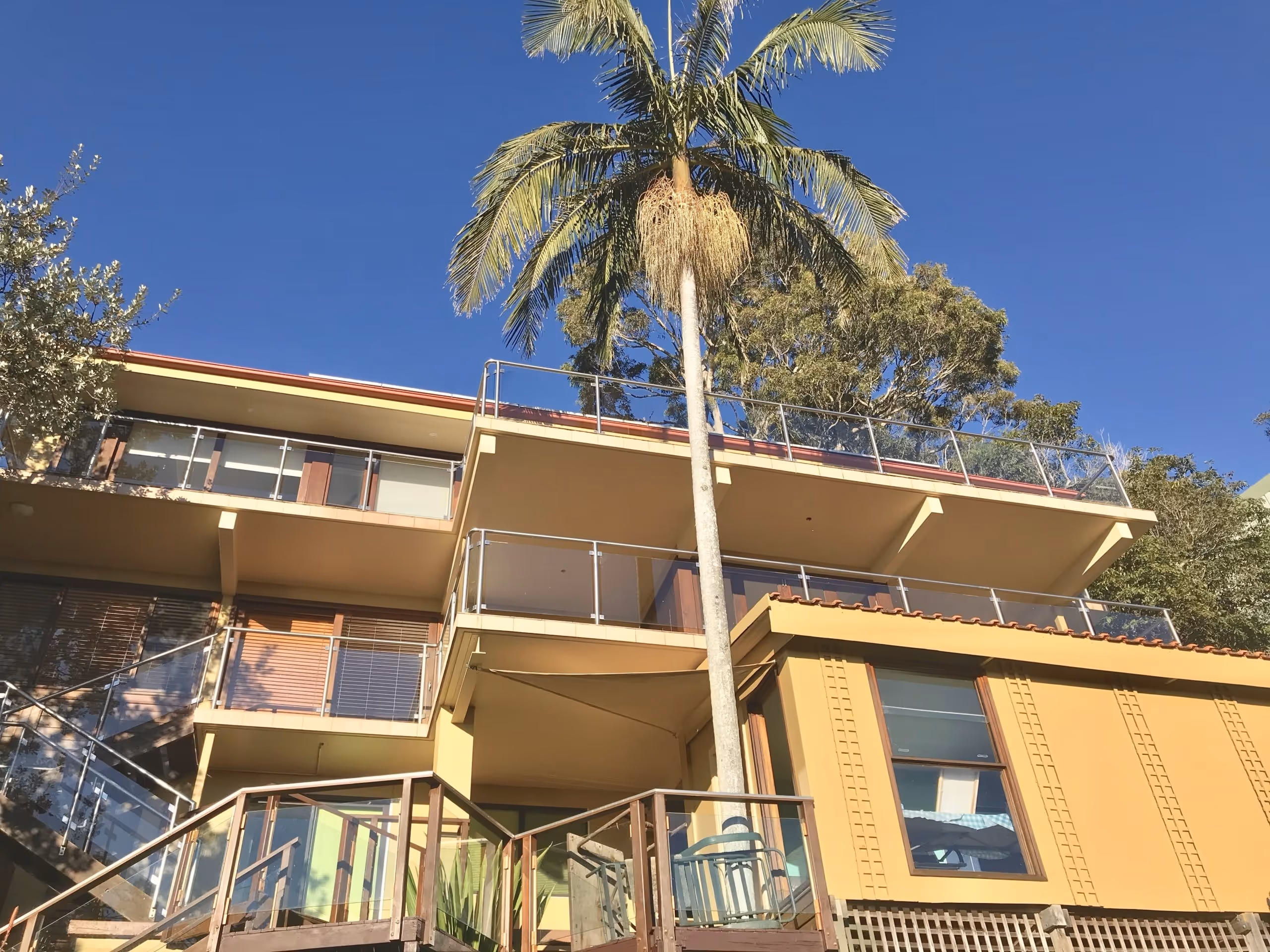 Modern multi-story yellow building with glass balconies and a tall palm tree in front under a clear blue sky.