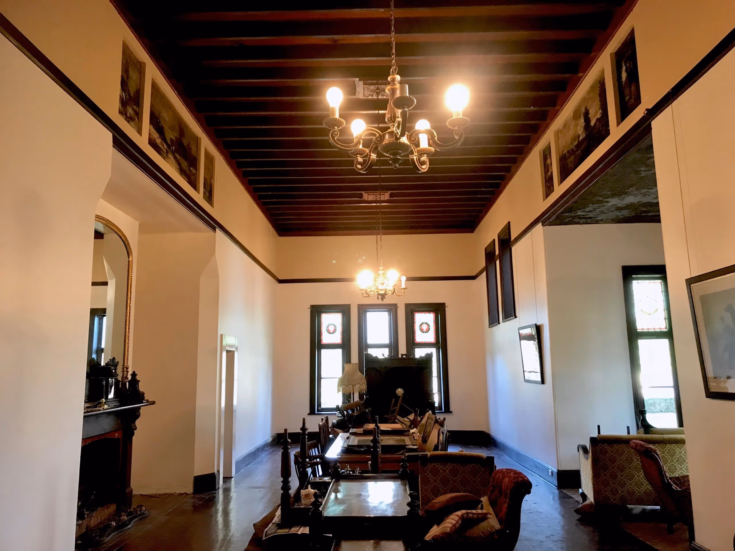 Interior of a vintage room with wooden ceiling beams, chandeliers, a fireplace, and antique furniture including chairs and tables.