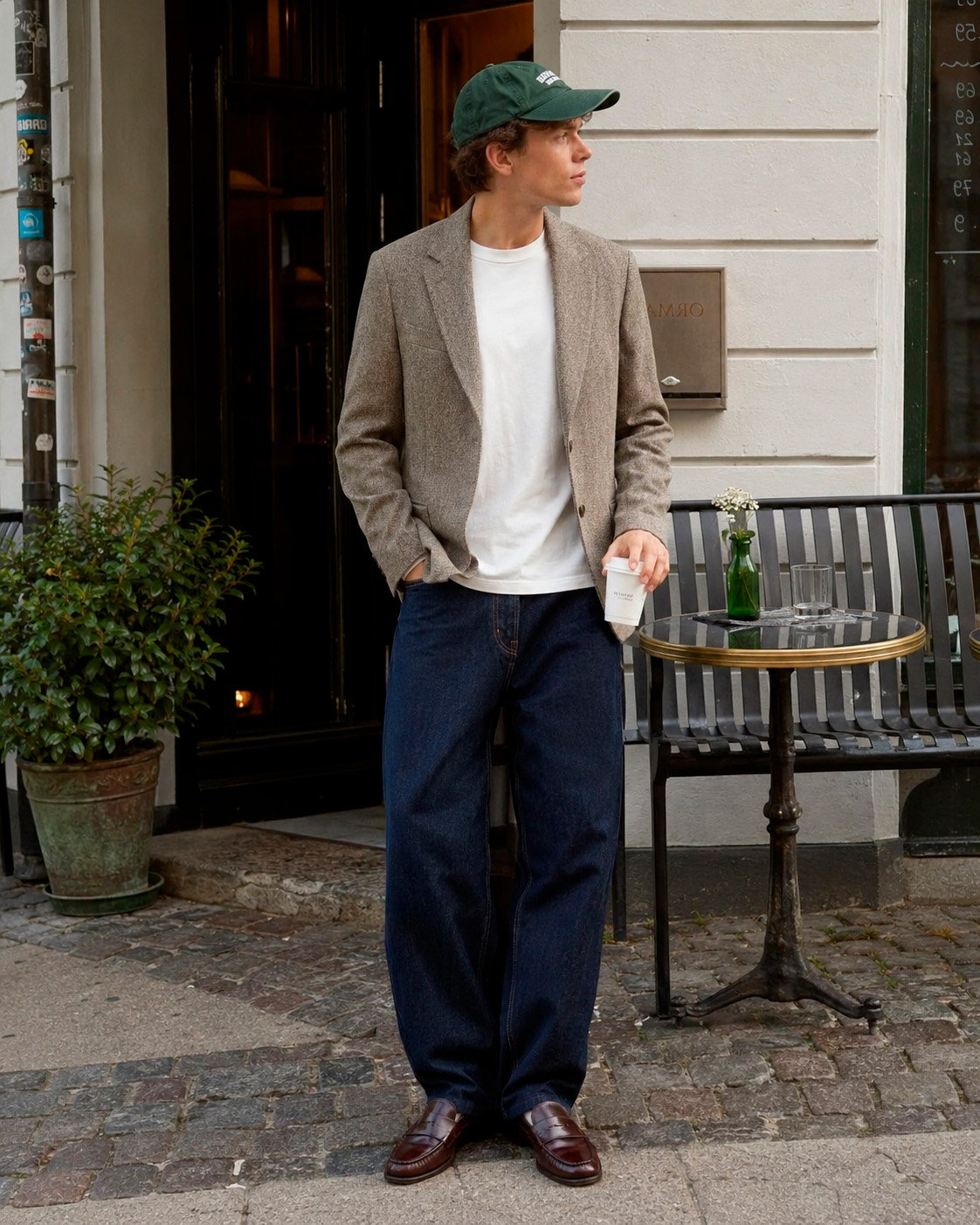 Young man wearing a textured taupe blazer, white T-shirt, wide dark jeans, green cap, and leather loafers outside a Copenhagen cafe.