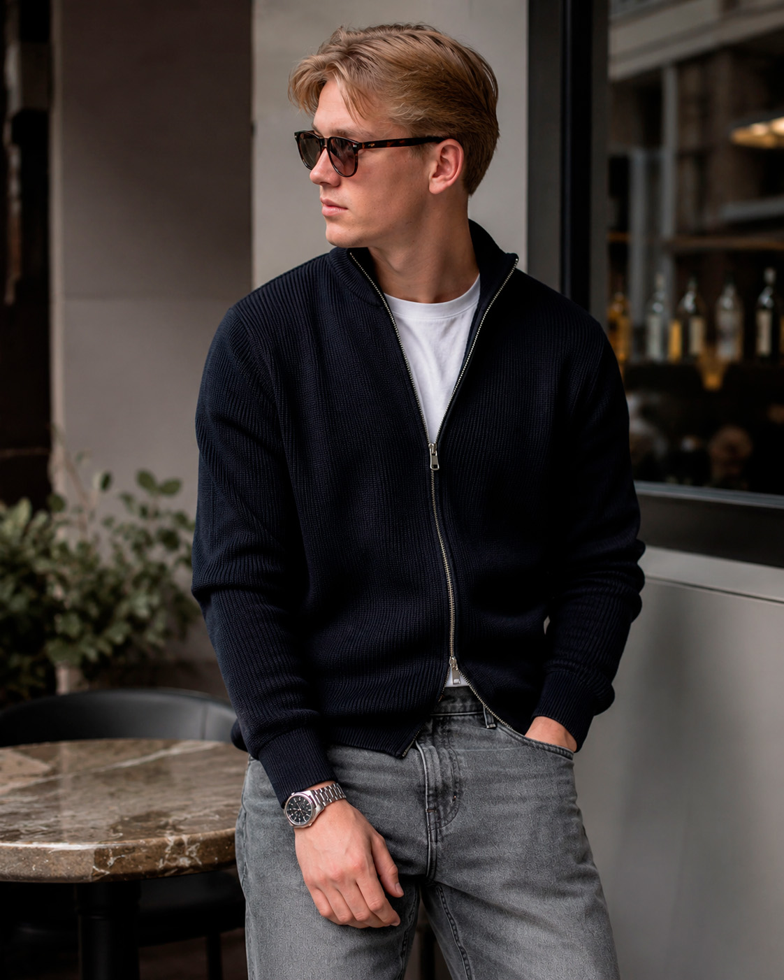 Young man in a dark zip knit, white T-shirt, grey jeans, sunglasses and a watch, styled in refined Grisch fashion.