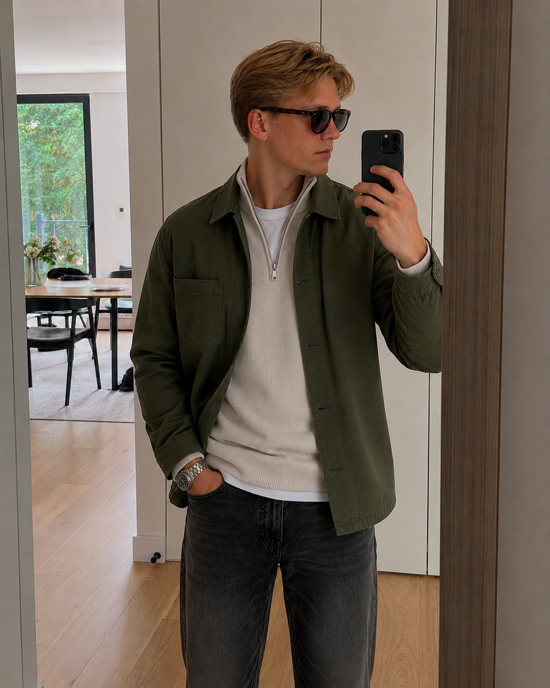 Mirror selfie of a young man wearing a dark quilted vest, zip knit, white T-shirt and grey jeans in a modern Grisch outfit.