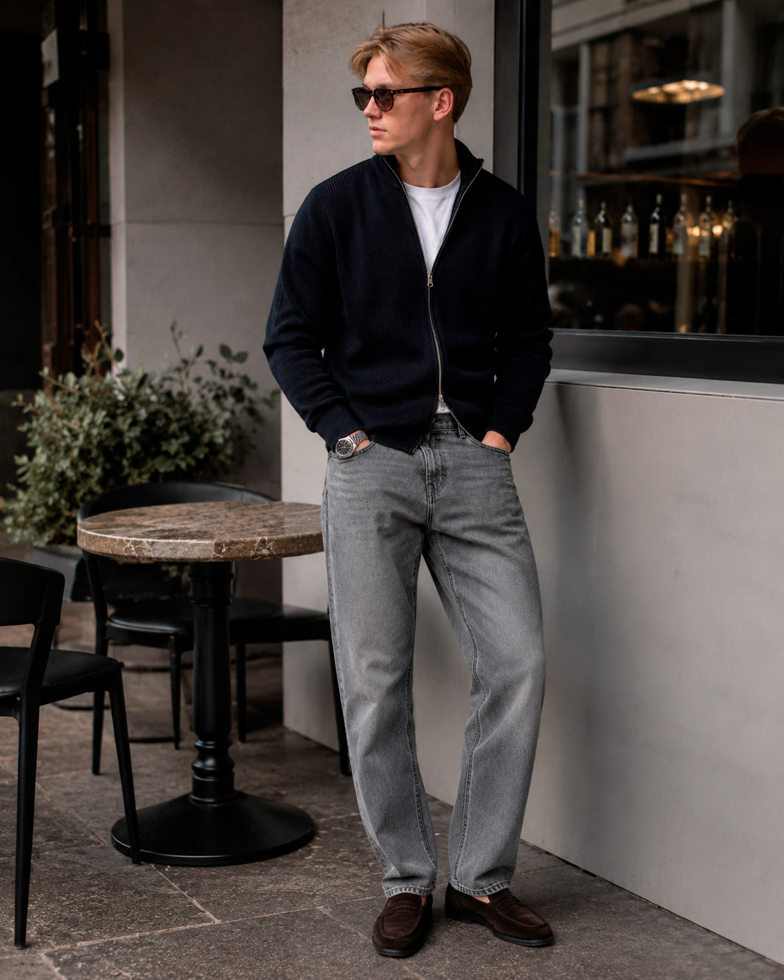 Young man standing outside a café in a dark zip knit, grey jeans, sunglasses and loafers, showing a polished Grisch style outfit.