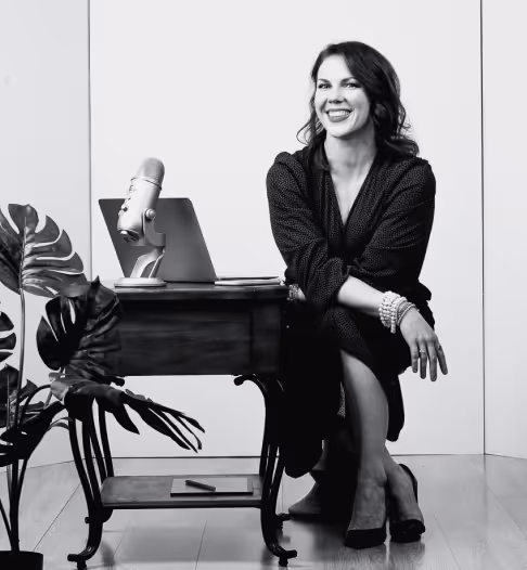 Black and white photo of a smiling woman sitting beside a small wooden table with a laptop and microphone, next to a large leafy plant.
