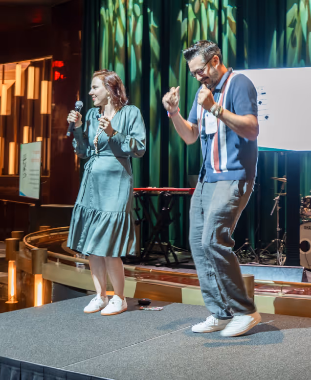 A woman in a green dress holding a microphone and a man in a navy blue shirt dancing on a stage with green curtains in the background.