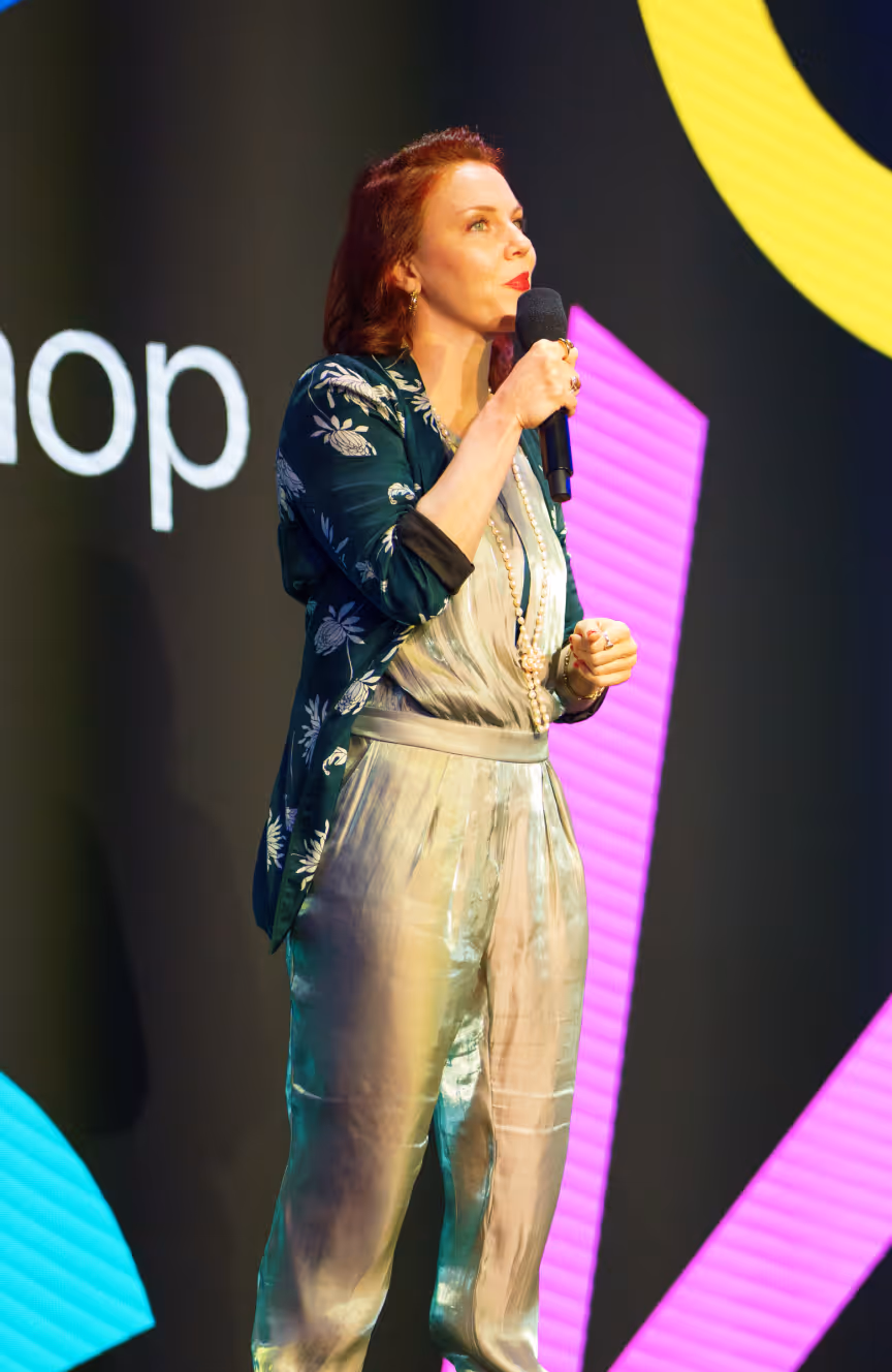 Woman with red hair speaking into a microphone on stage wearing a floral jacket and shiny pants.
