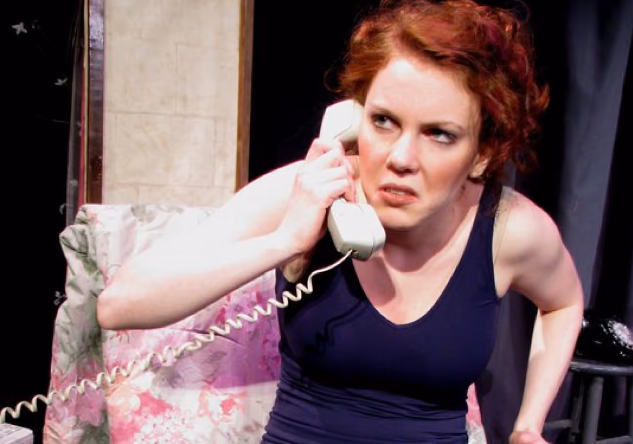 Woman with red hair and a black tank top looking angry while talking on a white corded telephone.
