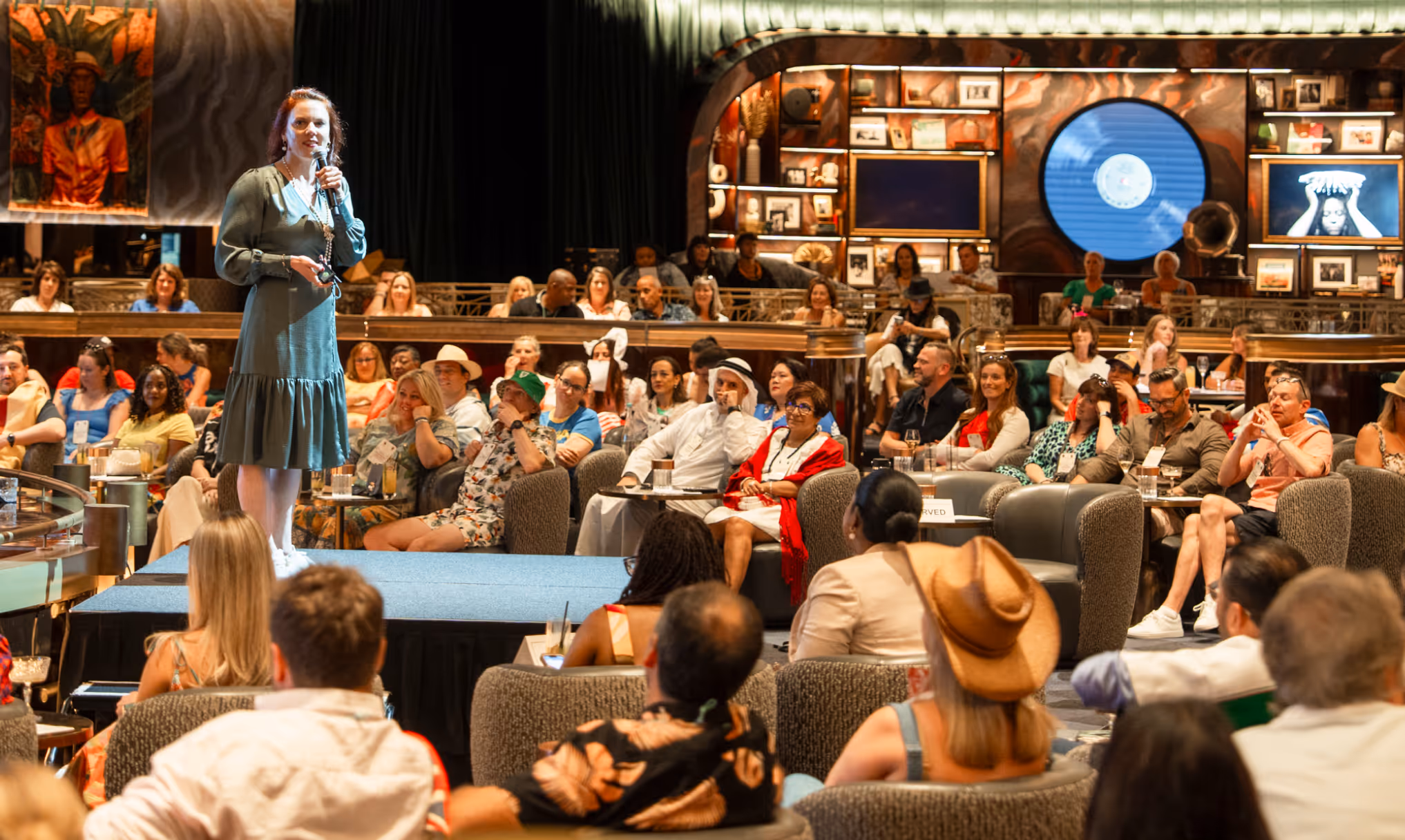 Woman in a green dress speaking on a stage in front of a seated audience in a lounge setting.