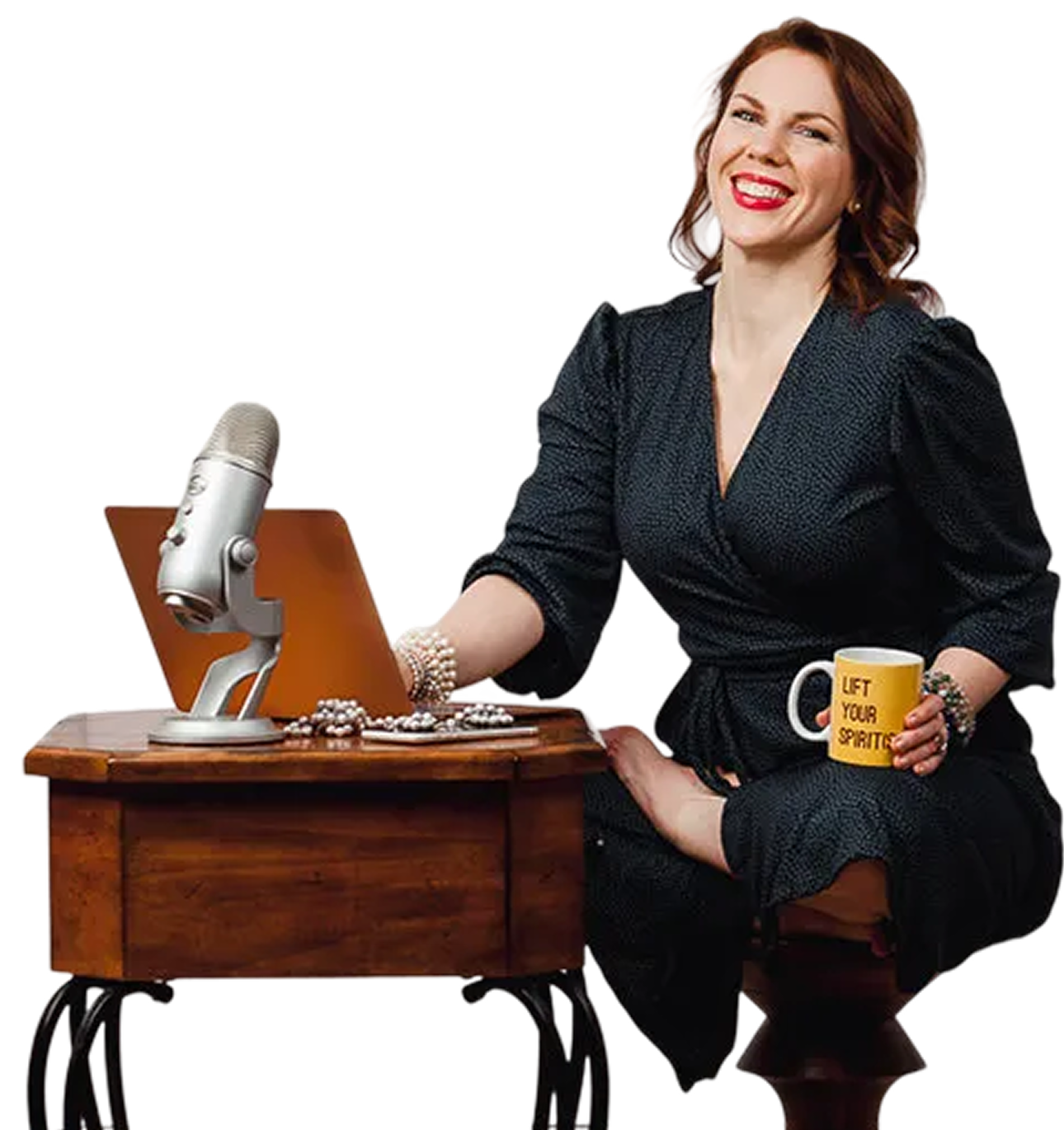Smiling woman in black dress sitting barefoot on a wooden stool, holding a yellow mug that says 'LIFT YOUR SPIRITS', with a laptop and microphone on a wooden table in front of her.
