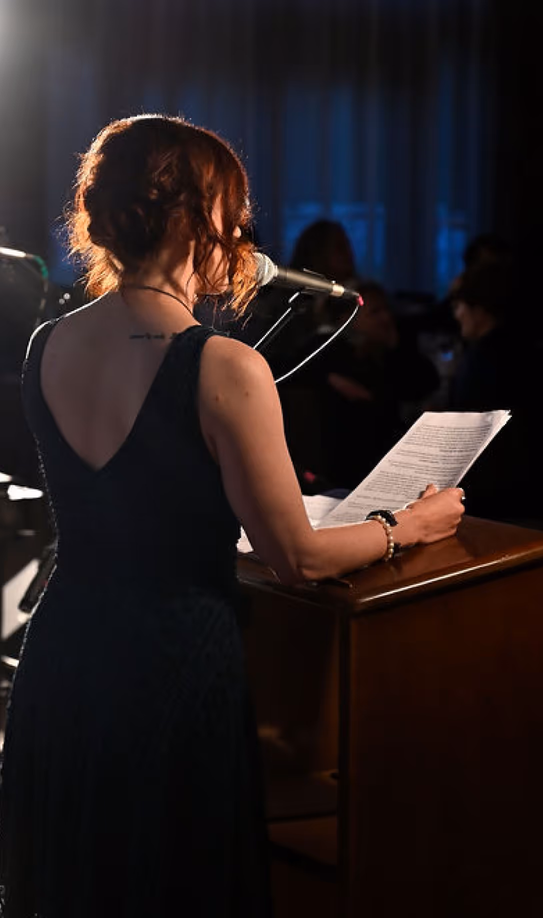 Woman with red hair in a dark dress speaking into a microphone while holding papers at a podium.