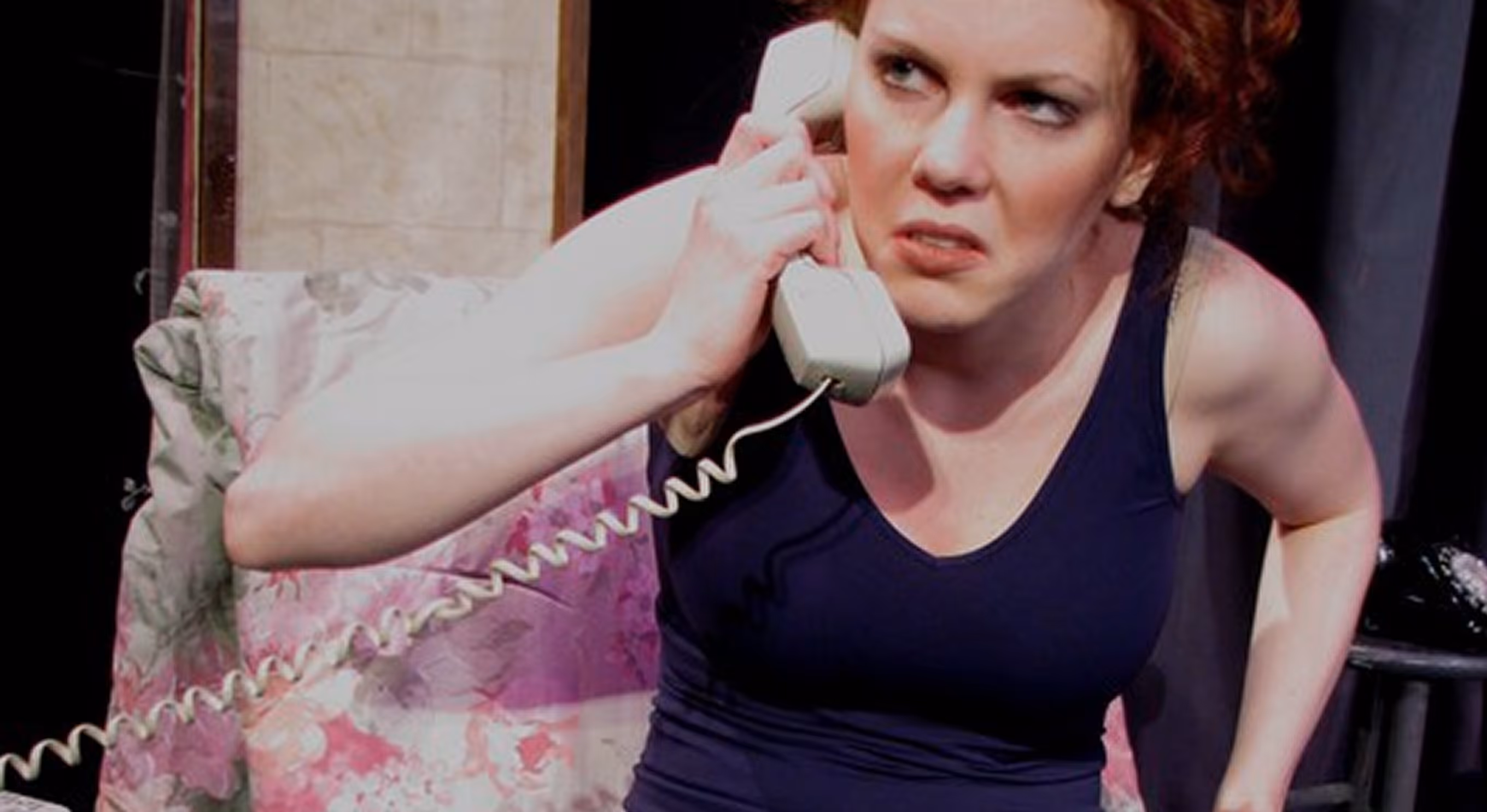Woman with red hair and a dark tank top holding a corded telephone receiver to her ear with a displeased expression.