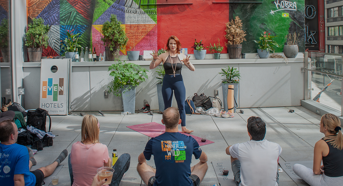 A woman instructing a group of people in a yoga or fitness class on an outdoor terrace with colorful murals and plants in the background.