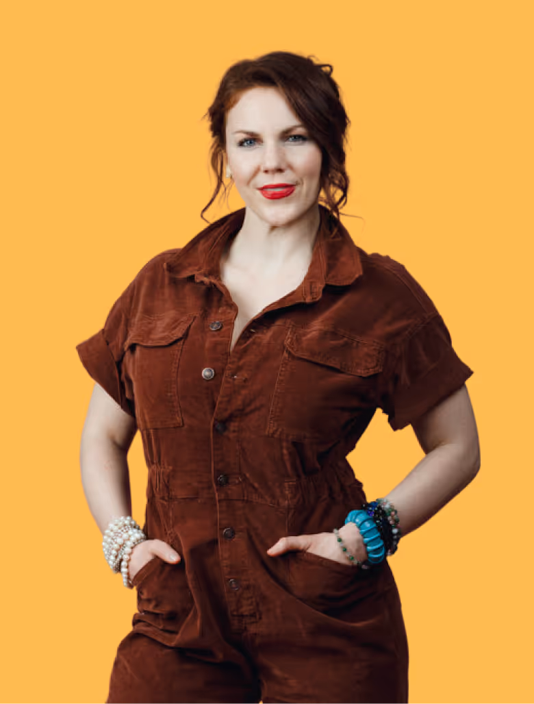 Smiling woman with red lipstick and red hair in a brown buttoned dress with hands in pockets against an orange background.