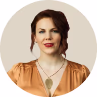 Portrait of a woman with red hair in an orange blouse and leaf pendant necklace, against a light beige circular background.