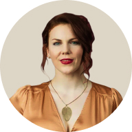 Portrait of a woman with red hair in an orange blouse and leaf pendant necklace, against a light beige circular background.