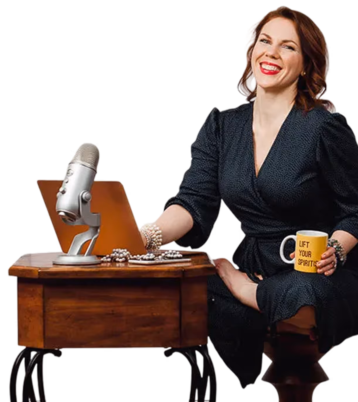 Smiling woman in black dress sitting cross-legged on a stool, holding a yellow mug that says 'LIFT YOUR SPIRITS,' next to a wooden table with a microphone, laptop, and pearl necklaces.