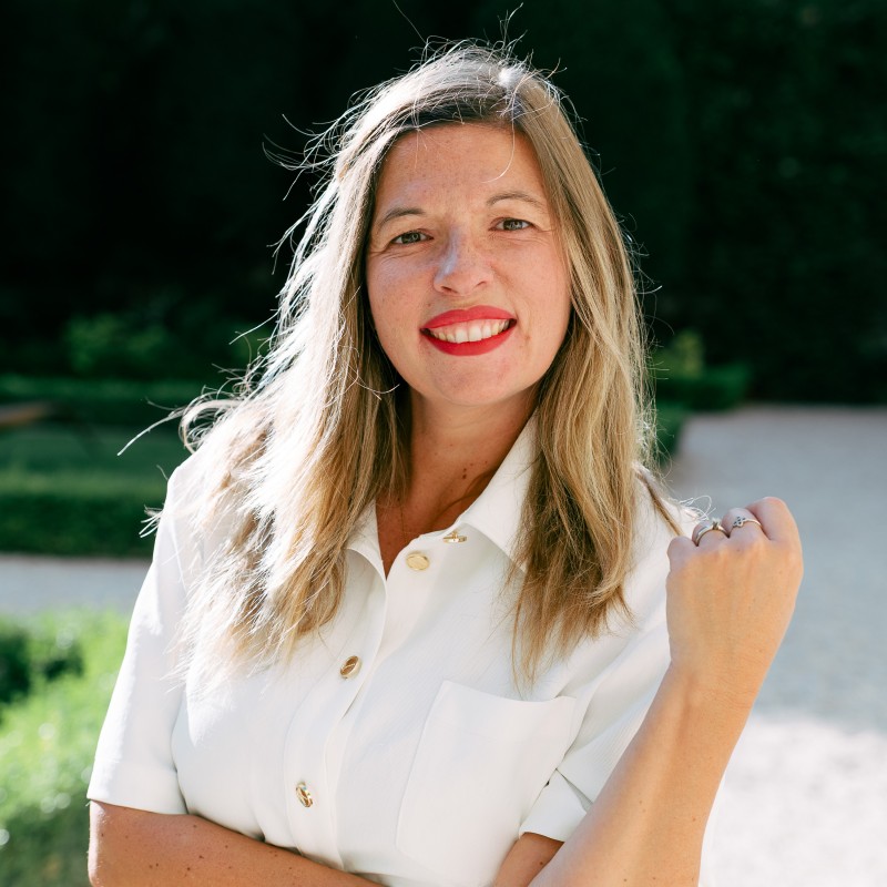 Femme aux longs cheveux blonds portant une chemise blanche souriant au soleil dans un jardin.
