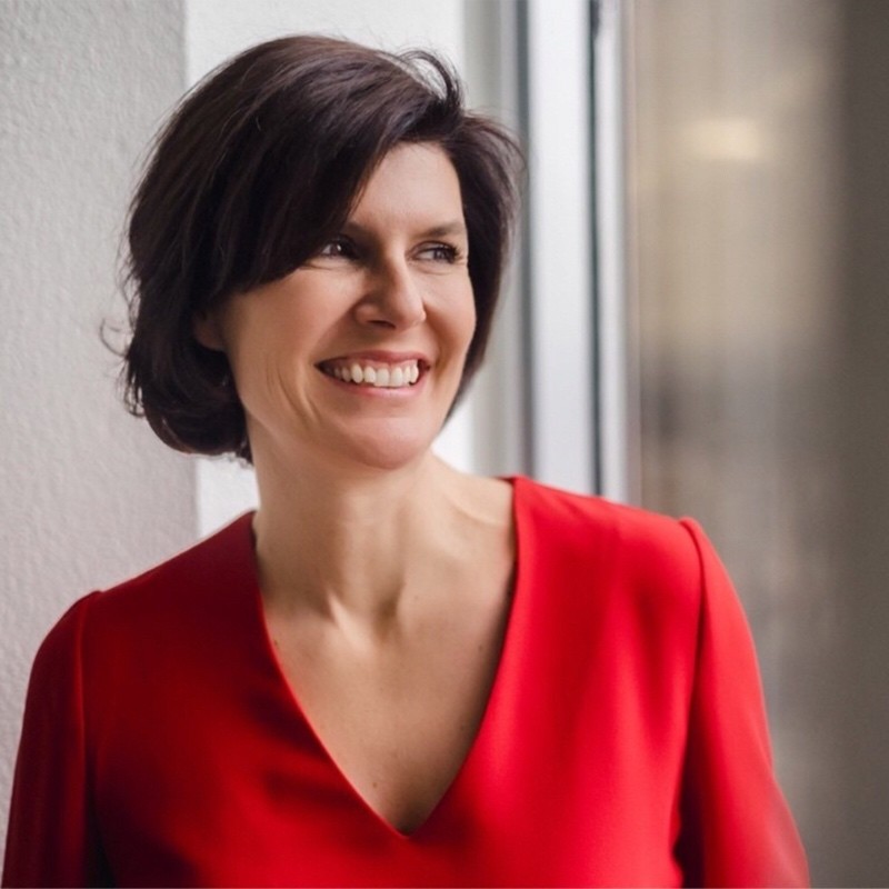 Femme souriante aux cheveux courts et bruns portant un haut rouge, regardant par une fenêtre.