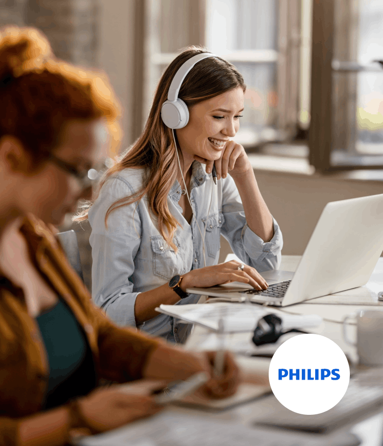 Une femme souriante portant un casque blanc utilise un ordinateur portable dans un bureau lumineux avec une autre personne floue au premier plan.