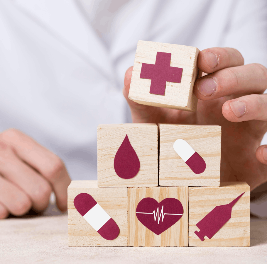 Une main empile des cubes en bois avec des icônes médicales, y compris une croix, une goutte de sang, des pilules, un cœur avec un électrocardiogramme et une seringue.