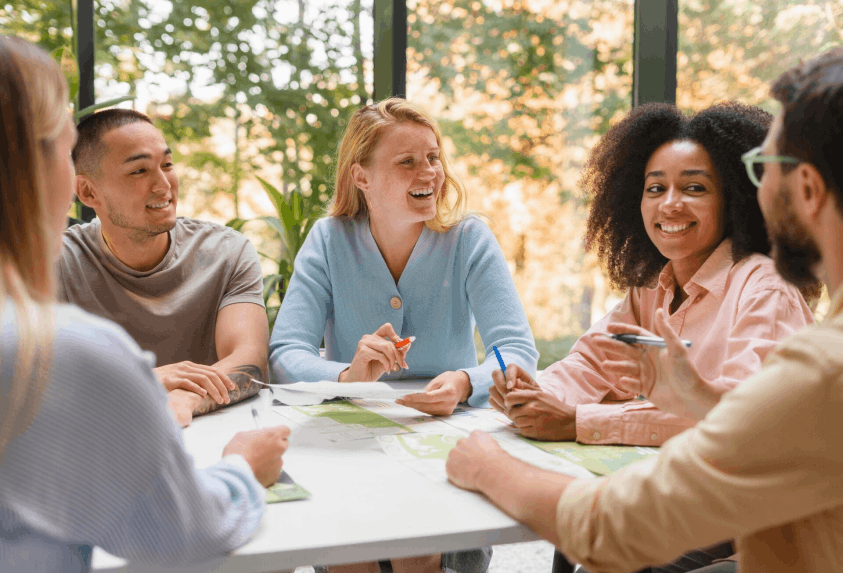 Cinq adultes de différentes origines discutent joyeusement autour d'une table blanche avec des documents, dans une salle lumineuse avec de grandes fenêtres.