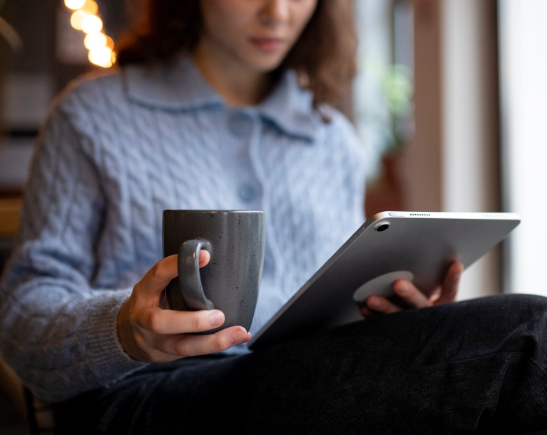 Une femme en pull gris tient une tasse et une tablette, assise près d'une fenêtre.