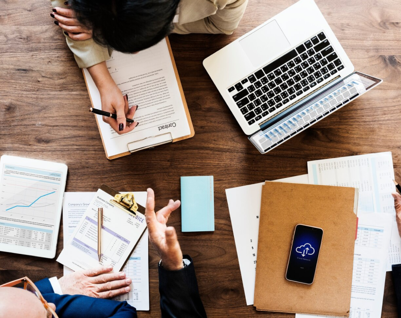 Vue aérienne d'une table en bois avec plusieurs personnes travaillant, une main prenant des notes sur une feuille de contact, un ordinateur portable ouvert, des documents financiers, et un smartphone affichant une icône de cloud.