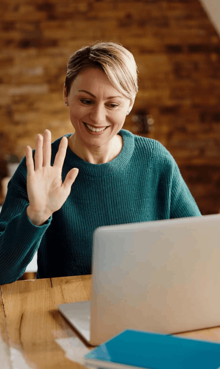 Une femme souriante en pull bleu saluant devant un ordinateur portable.