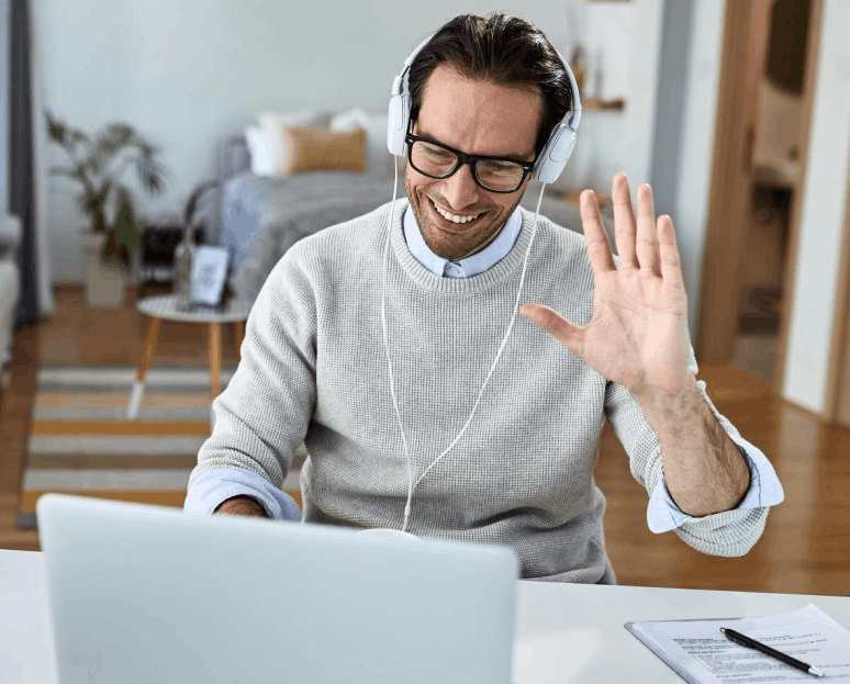 Un homme souriant portant des lunettes et un casque écoute sur un ordinateur portable et fait un geste de salutation de la main droite.