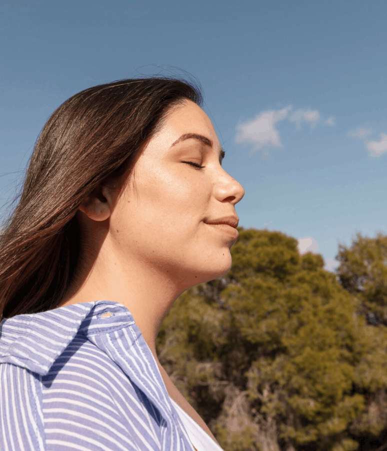 Une femme aux longs cheveux bruns, les yeux fermés, profite du soleil avec un léger sourire, devant un ciel bleu et des arbres verts.