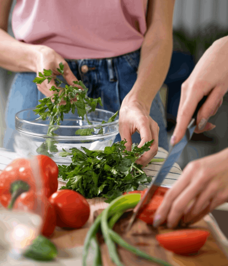 Deux personnes préparent une salade en éminçant des tomates et en plaçant du persil dans un bol en verre.
