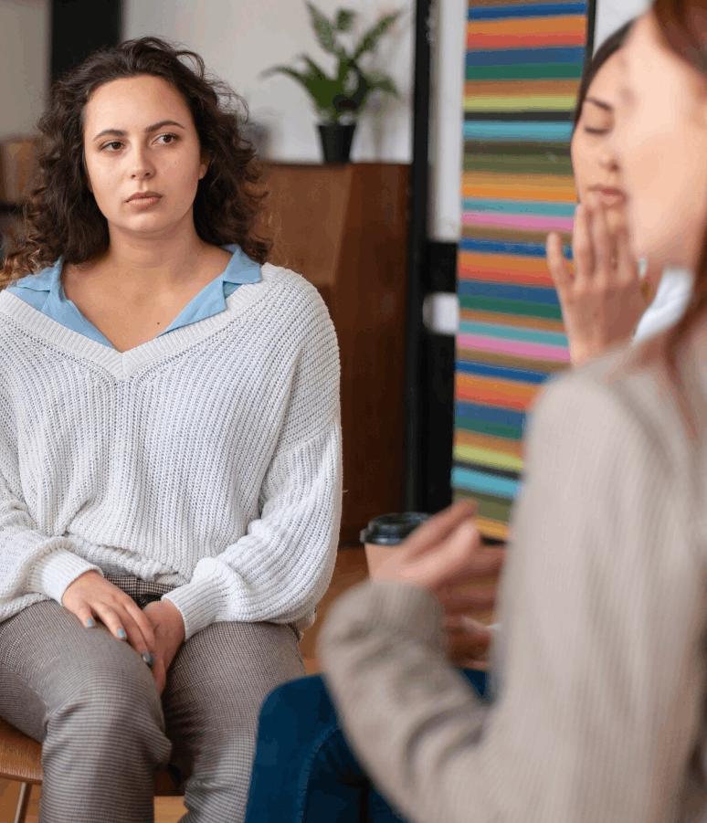 Une femme assise écoutant attentivement une autre personne parlant, dans une ambiance intérieure confortable.