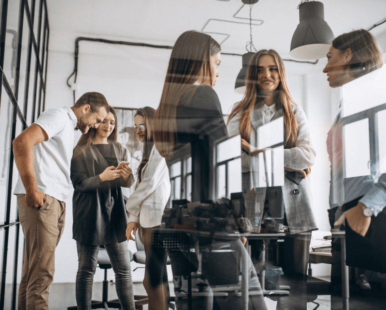 Un groupe de six jeunes professionnels discutant et regardant un téléphone dans un bureau moderne lumineux avec des murs blancs et des fenêtres larges.