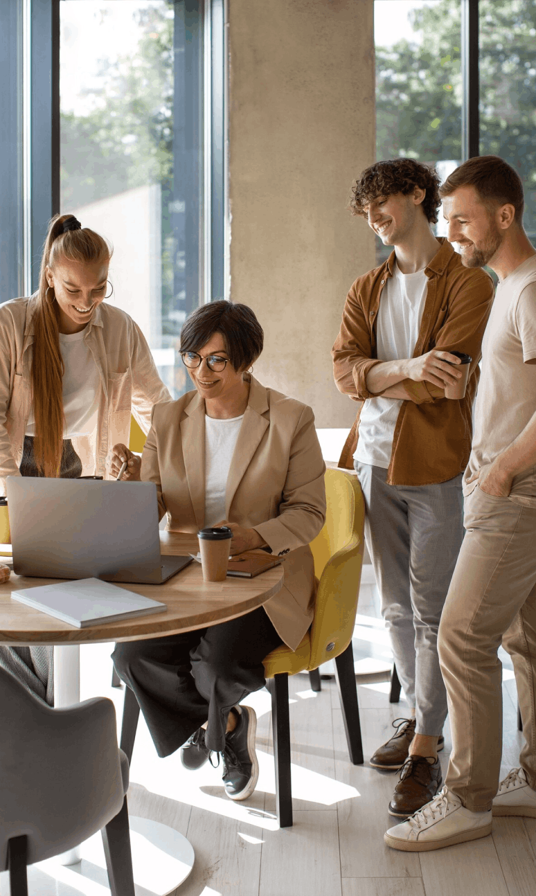 Quatre collègues souriants collaborent autour d'un ordinateur portable dans un bureau lumineux avec de grandes fenêtres.