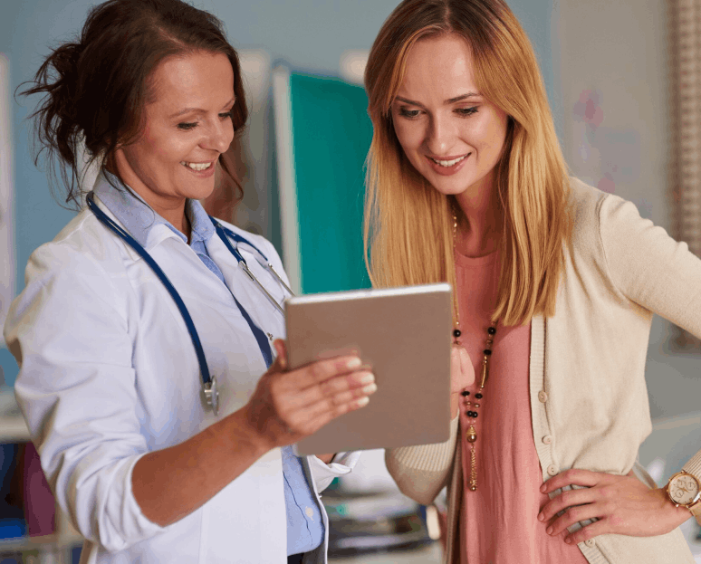 Une médecin souriante montre quelque chose sur une tablette à une patiente blonde qui regarde avec intérêt.