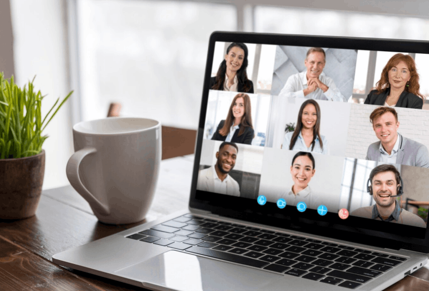 Ordinateur portable ouvert sur une réunion vidéo avec neuf participants souriants, une tasse blanche et une plante verte à côté.