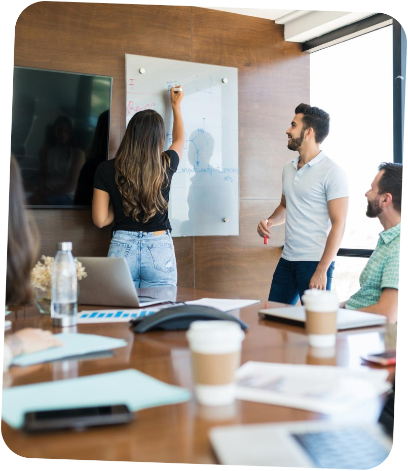 Femme écrivant sur un tableau blanc en verre pendant que deux hommes l'écoutent dans une salle de réunion lumineuse.