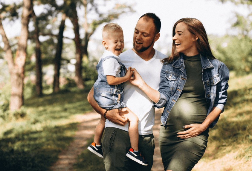 Famille heureuse en promenade dans un parc, un homme tenant un enfant riant, une femme enceinte souriante à côté.