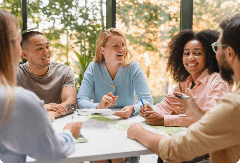Un groupe de cinq personnes souriantes discutant autour d'une table avec des plans sur une grande fenêtre en arrière-plan.