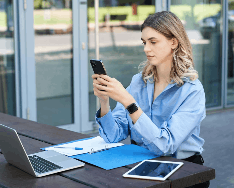 Jeune femme assise à l'extérieur, utilisant un smartphone avec un ordinateur portable, une tablette et des dossiers sur la table.